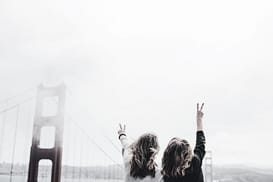 two women on the background of a large bridge show someone with their hands a sign of peace